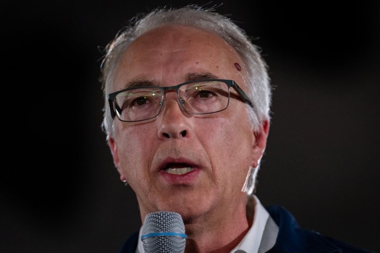 A man with white hair and glasses speaks into a microphone.