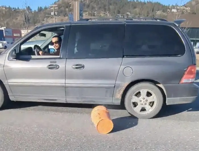 A man with sunglasses driving a grey van after throwing stuff out the window, and an orange bucket can be seen on the road.