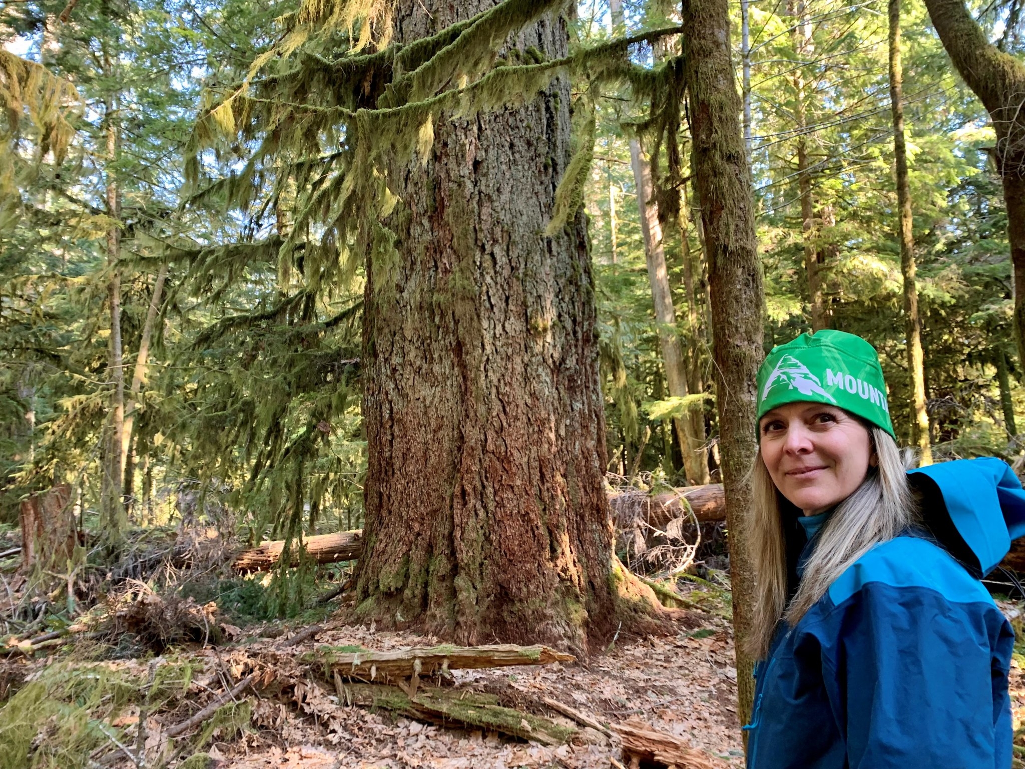 A woman smiles in the foreground with an old growth tree and forest in the background.
