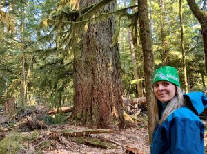 A woman smiles in the foreground with an old growth tree and forest in the background.