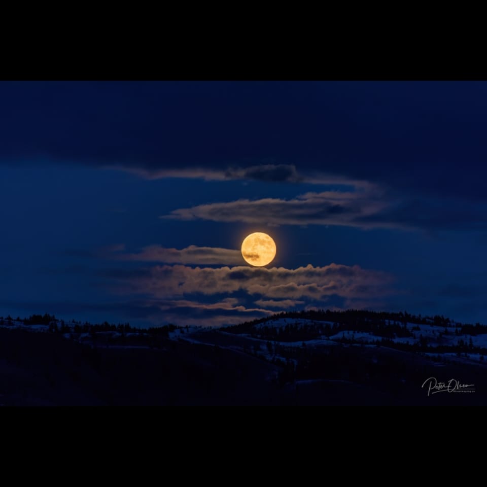 A full moon shines from behind whisps of clouds in a dark blue night sky.