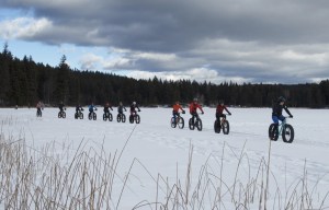 A row of riders on fat bikes travel on an open, snowy landscape.