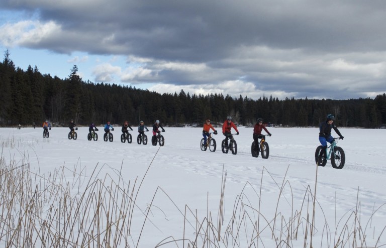 A row of riders on fat bikes travel on an open, snowy landscape.