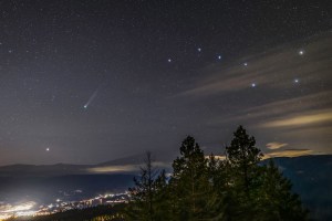A night sky full of stars, the big dipper and a comet are seen over the distant lights of a town.