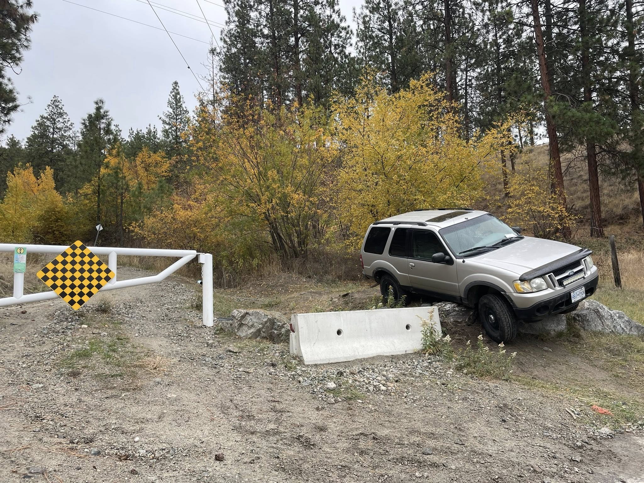 A silver Ford Explorer is high-centred on a rock on the side of a dirt road in the forest.