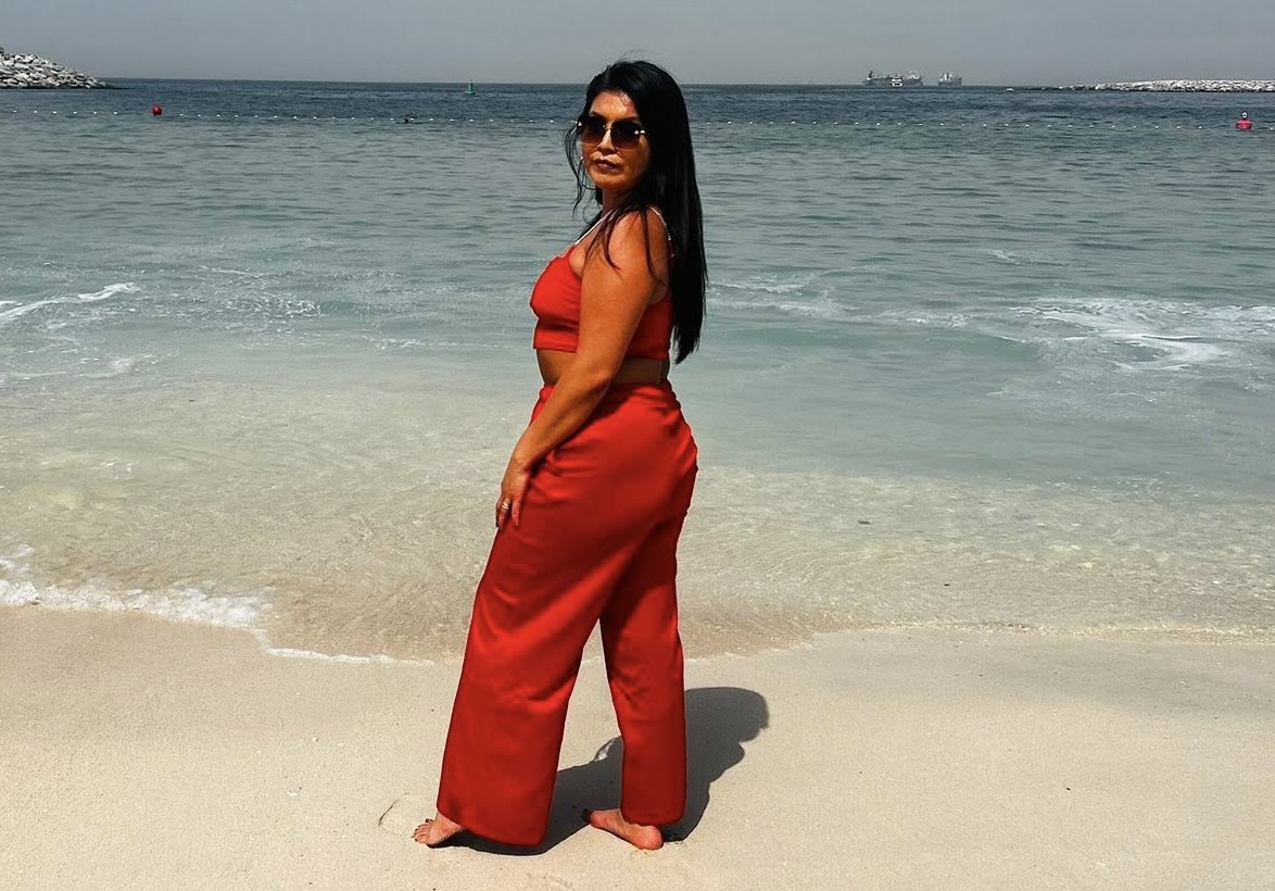 A woman stands in a red outfit on a sunny beach.