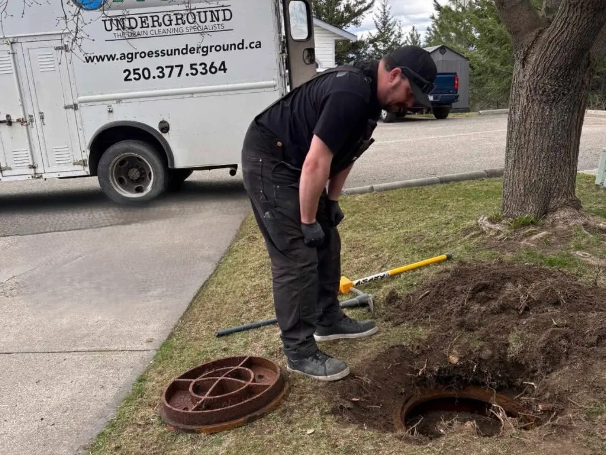 A worker looks into a septic tank on a lawn with his work vehicle in the background.