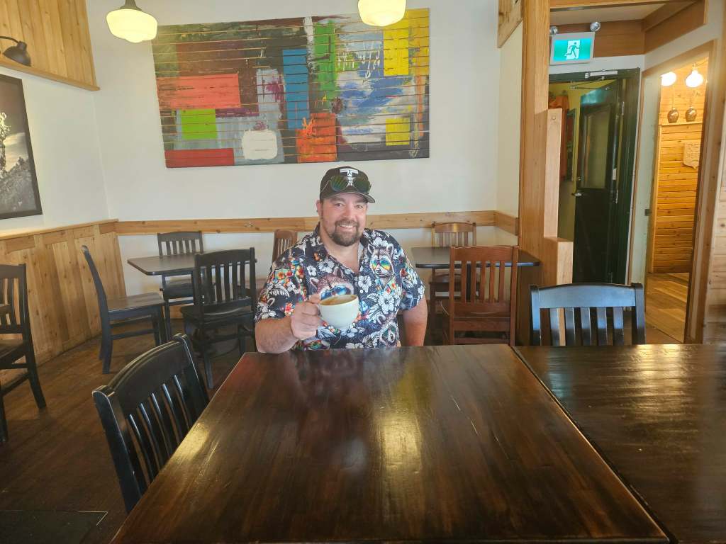 A man smiles from his seat behind a table in a coffee shop.
