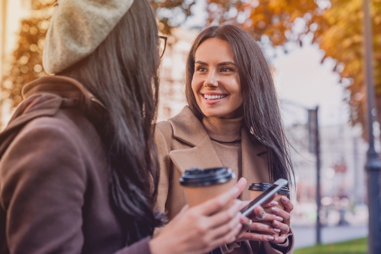 Young female friends talking with each other with hot beverages and a cellphone.