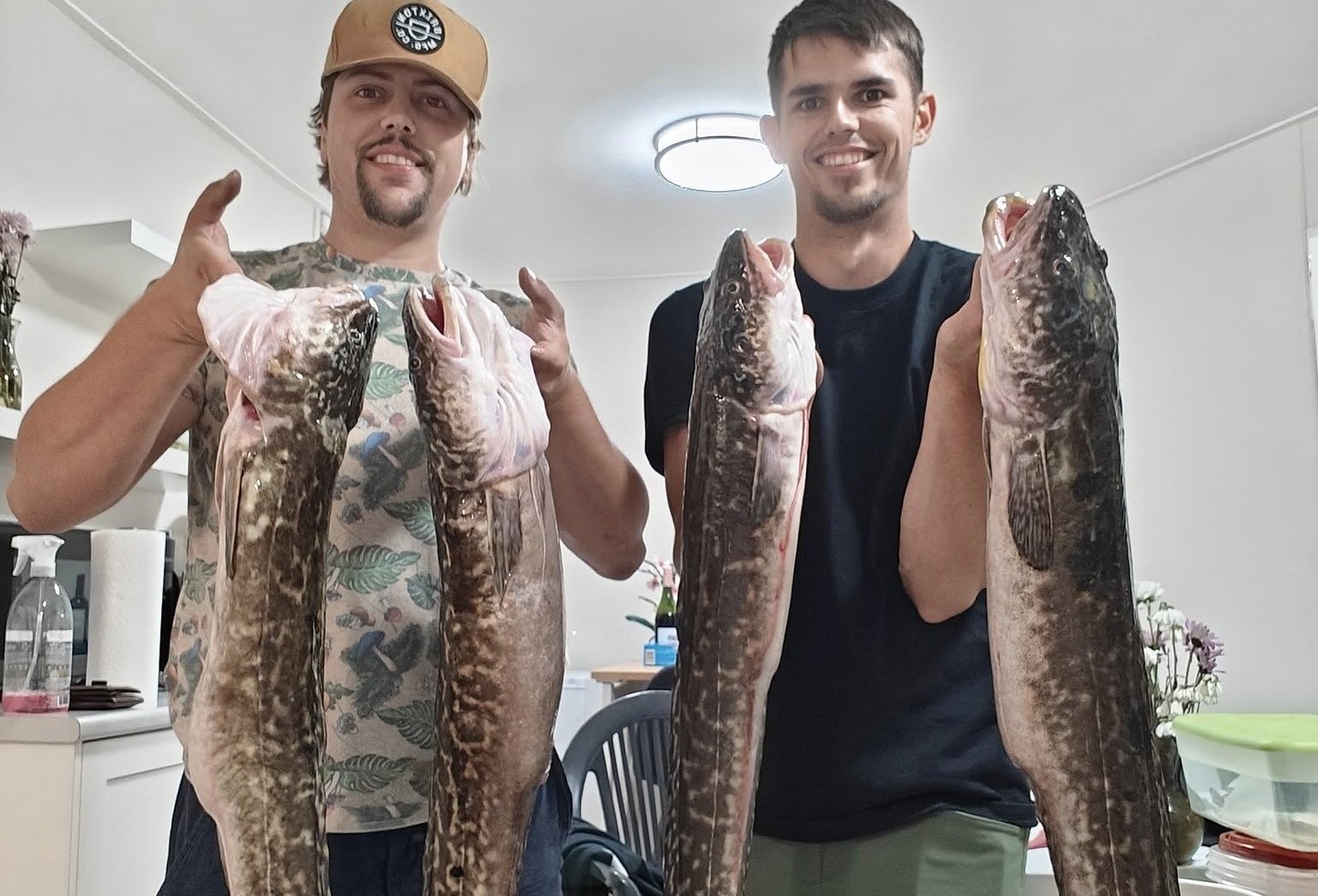 Two smiling men each hold up two long fish called burbot.
