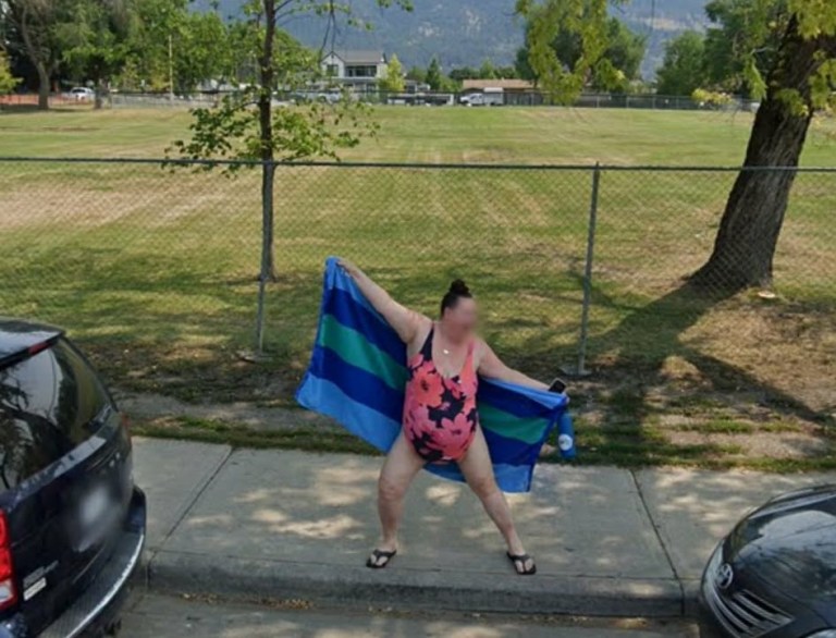 A woman in a bathing suite is standing up stretching a beach towel out behind her back.