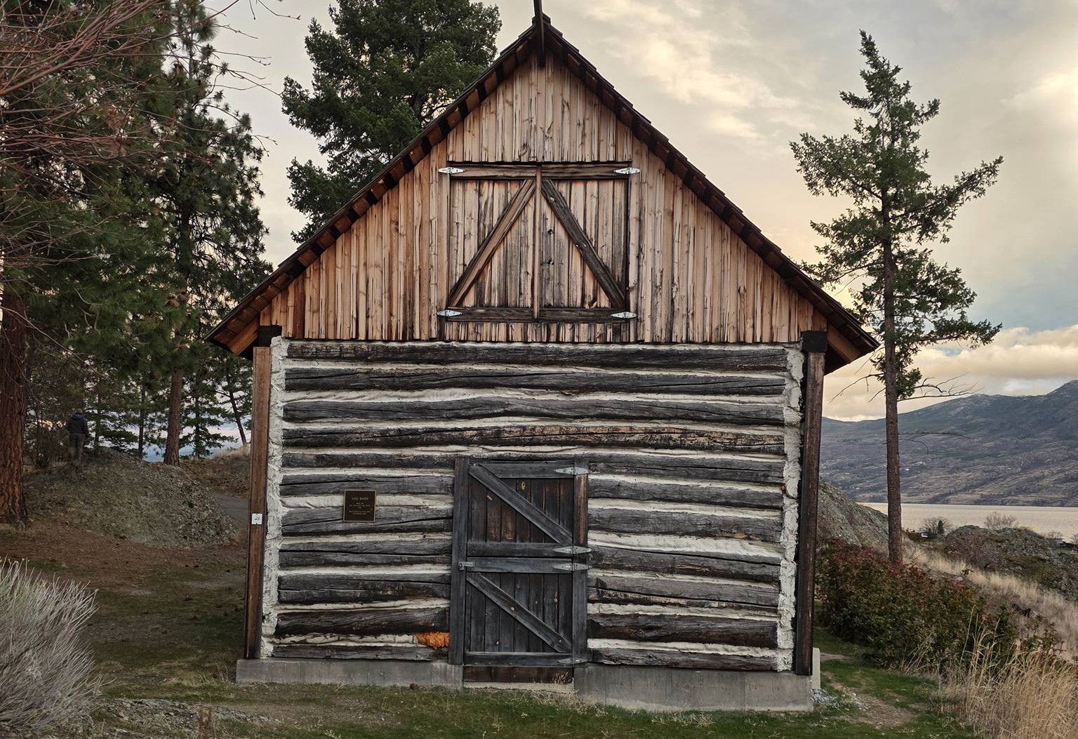 An old log barn is backed by a few conifer trees.