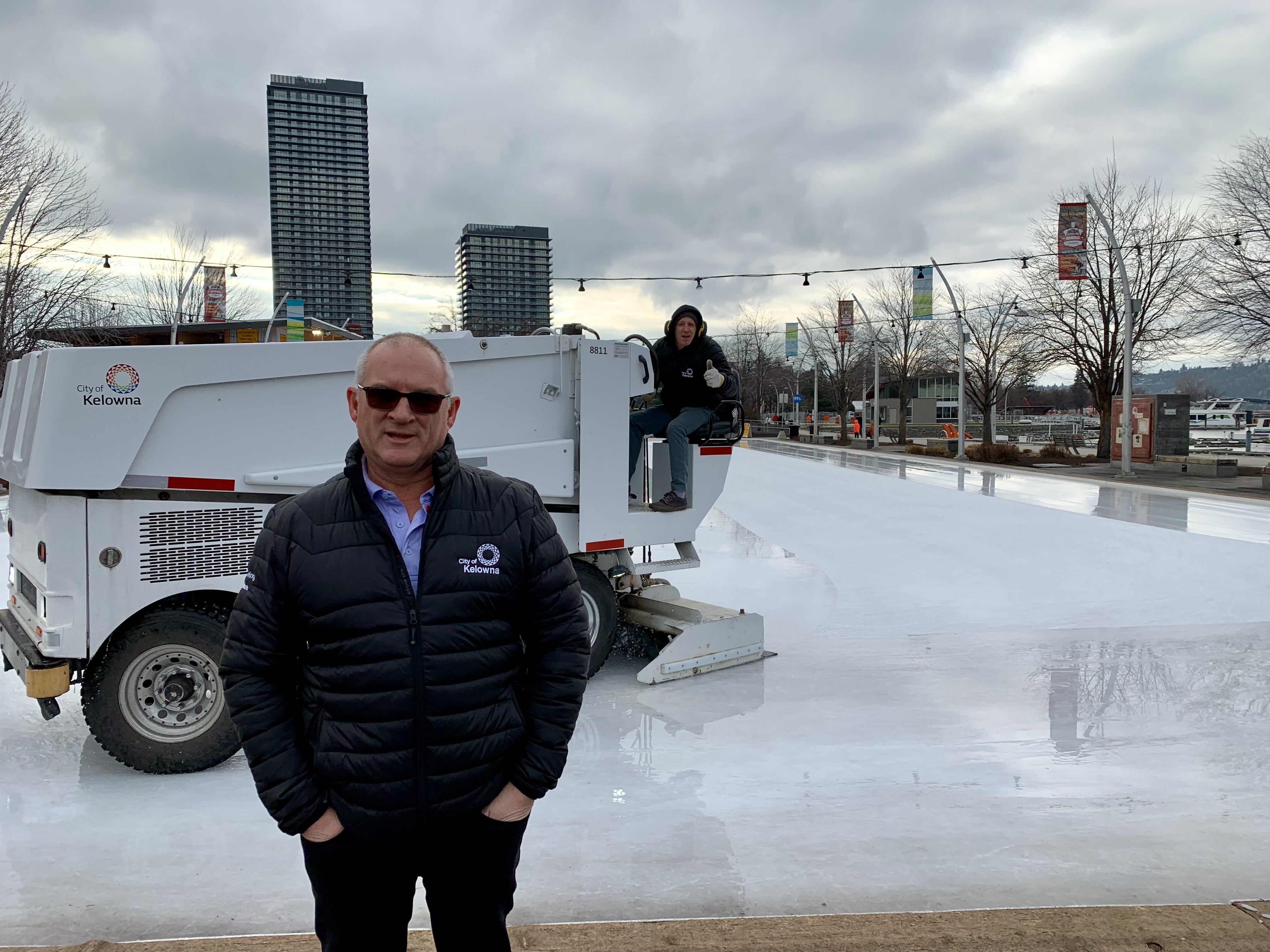 A man stands in front of an outdoor rink. Another rides a Zamboni.