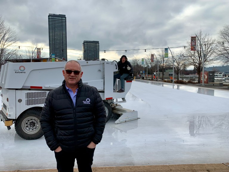 A man stands in front of an outdoor rink. Another rides a Zamboni.