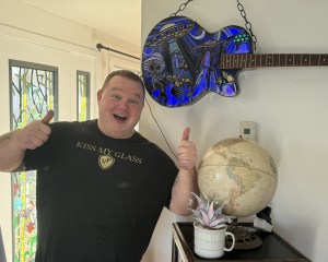A man standing in front of a stained glass guitar art piece holds two thumbs up.