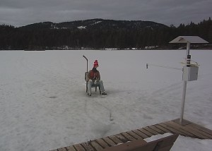 A mannequin of a man dressed in winter gear is set up on a frozen lake appearing to ice fish.