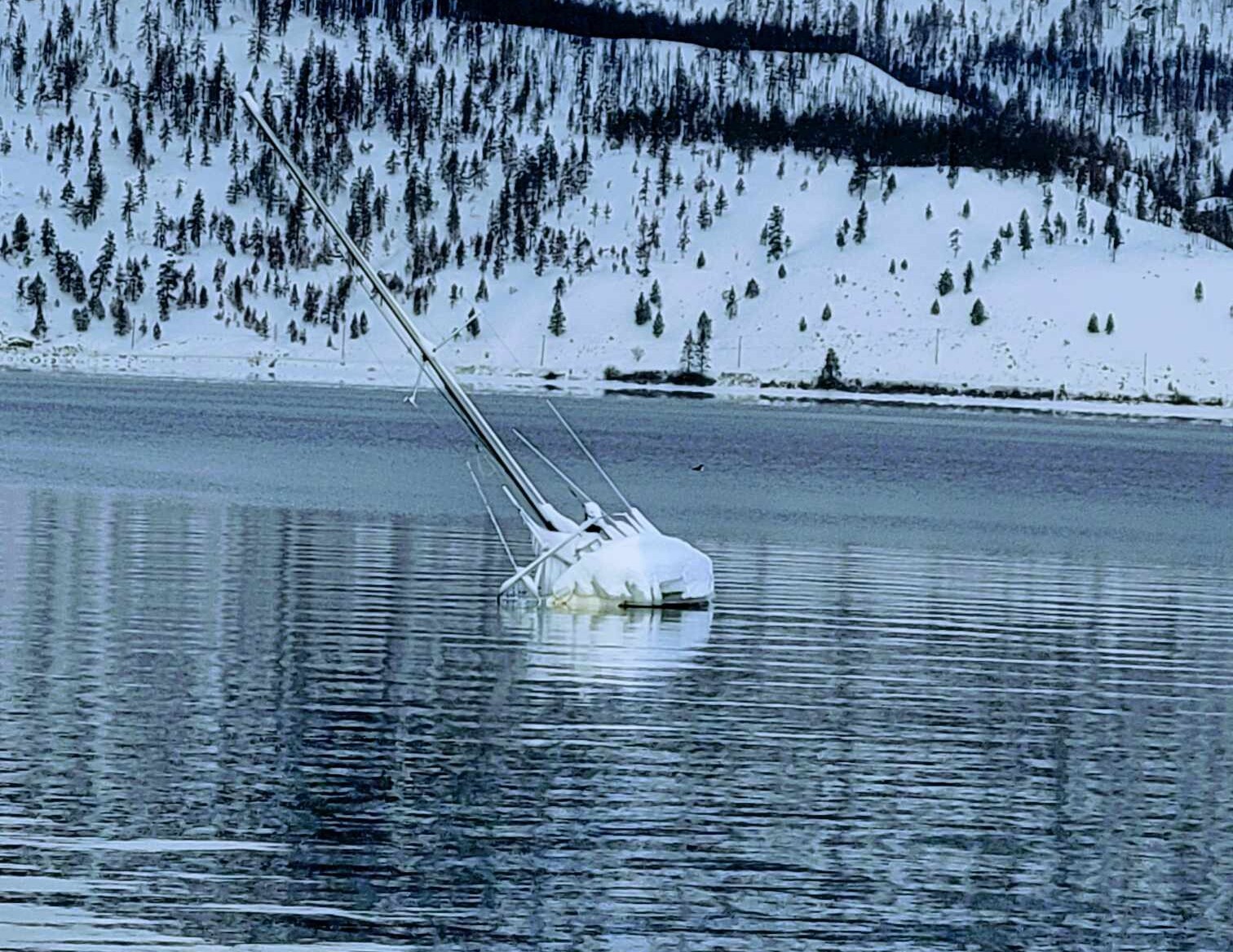 A sailboat is partially submerged on Okanagan Lake with snow covered hills in the background.