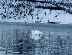 A sailboat is partially submerged on Okanagan Lake with snow covered hills in the background.