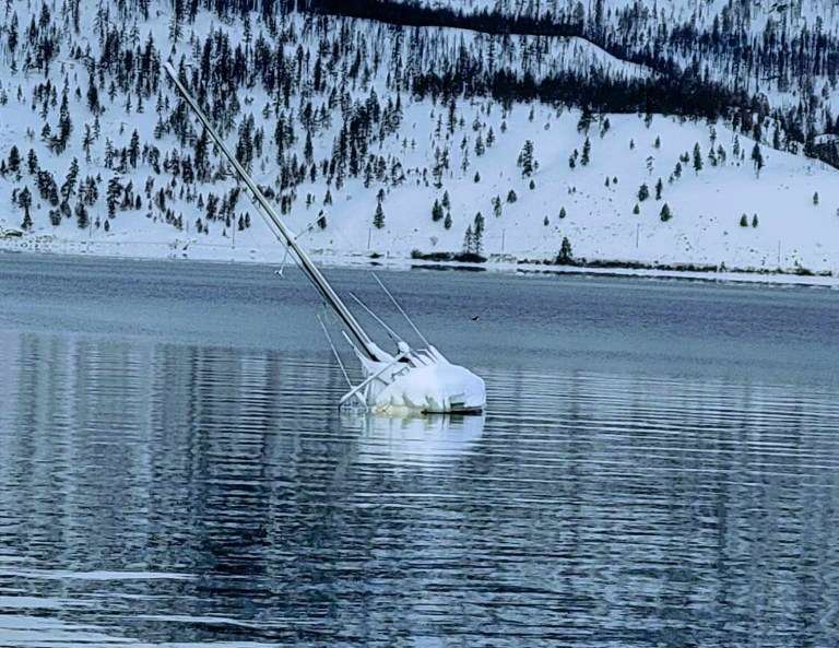 A sailboat is partially submerged on Okanagan Lake with snow covered hills in the background.