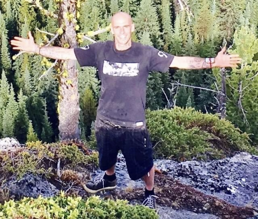 A man stands in a wooded area with his arms outstretched.