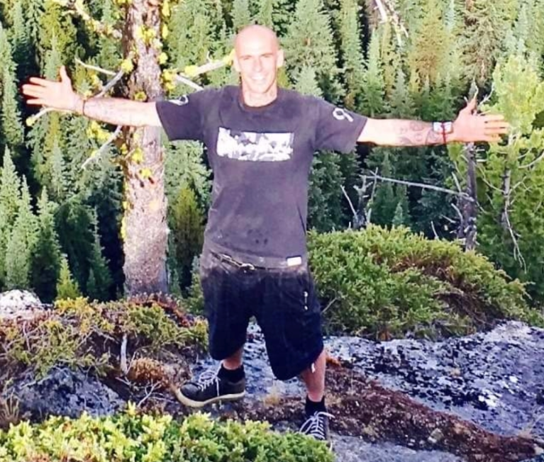 A man stands in a wooded area with his arms outstretched.