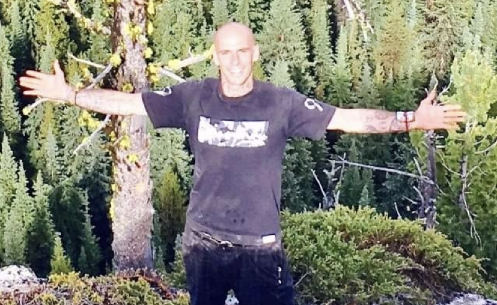 A man stands in a wooded area with his arms outstretched.