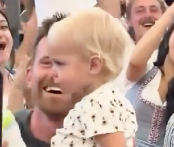 A baby standing with a crowd of people cheering in the background.