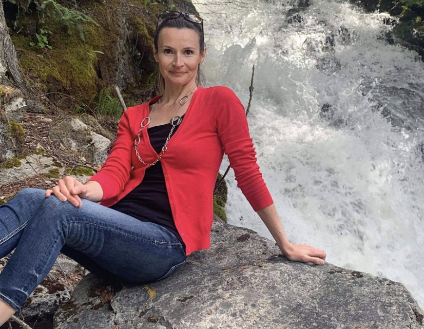 A woman wearing a black top, red jacket and jeans sits on a rock by a waterfall.