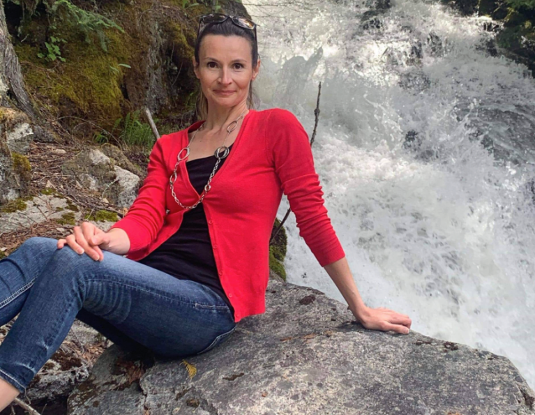 A woman wearing a black top, red jacket and jeans sits on a rock by a waterfall.