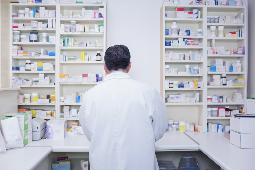 A pharmacist in a white coat stands in front of shelves full of drugs.