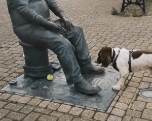 A dog looking at a ball underneath a statue of a man sitting.