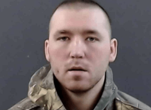 A mugshot of a man with a buzzcut in a camouflage sweater.