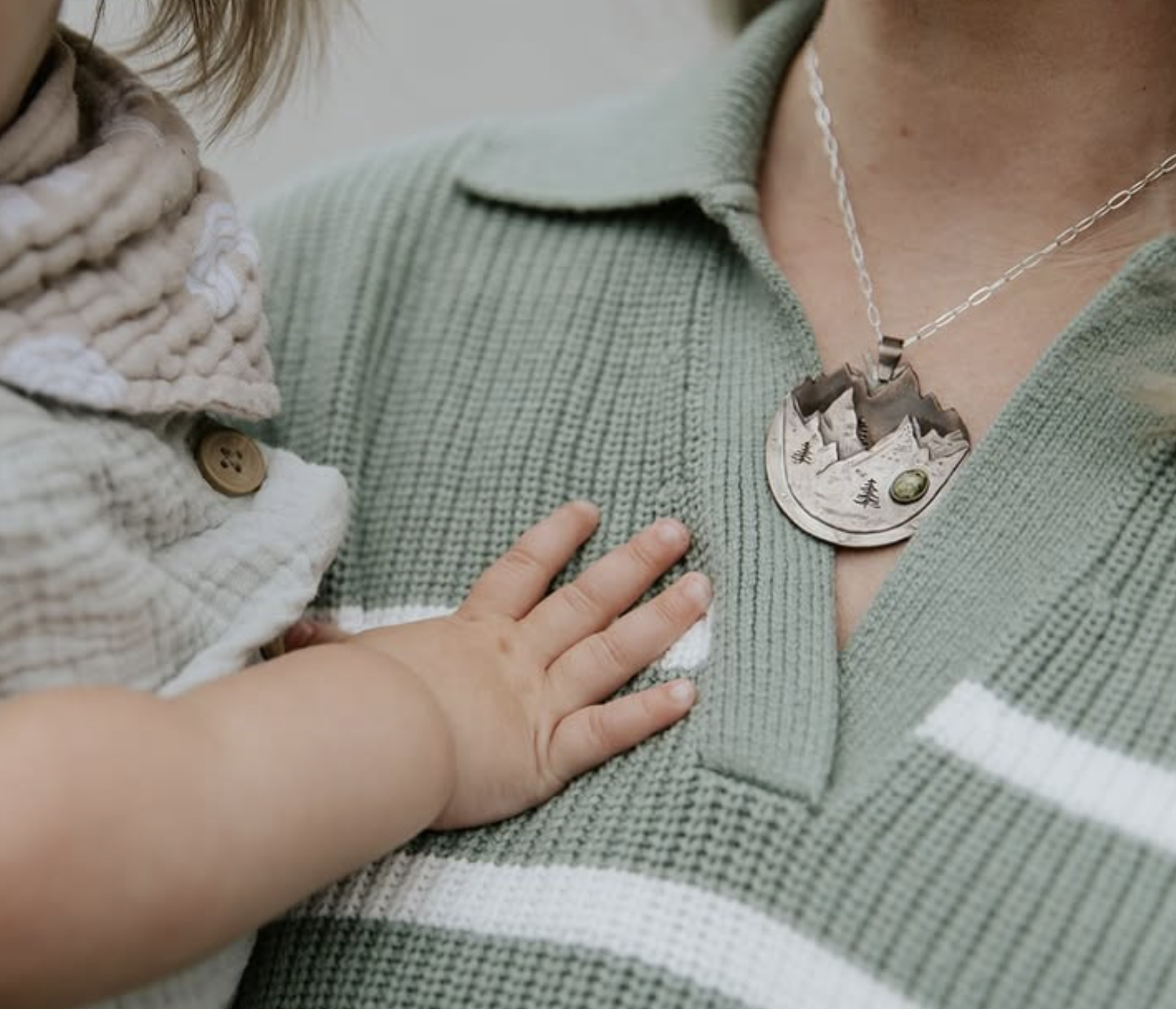A woman holding a toddler while wearing a necklace of mountains.