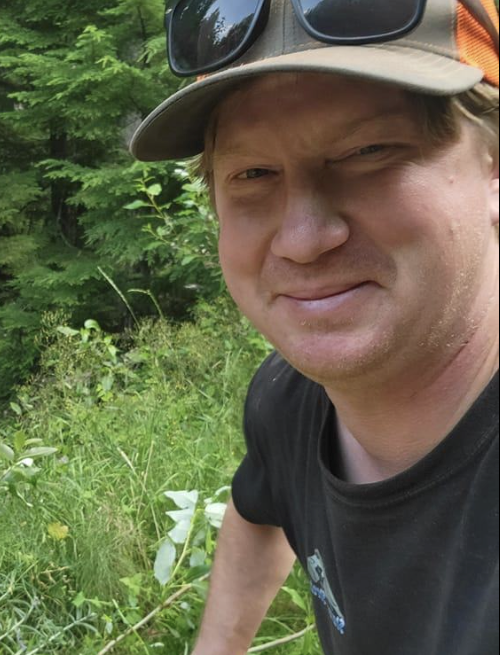 A man smile at the camera with greenery behind him.