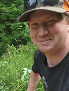 A man smile at the camera with greenery behind him.
