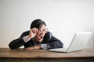 A person in a suit wearing glasses yells at a laptop computer on a desk.