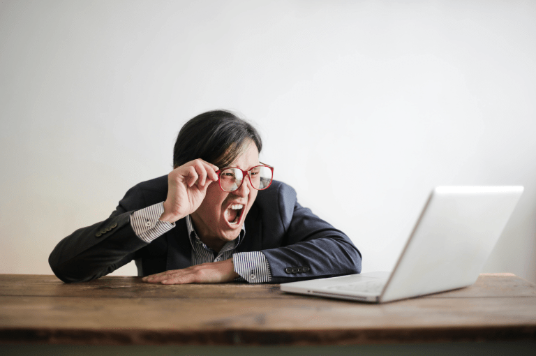 A person in a suit wearing glasses yells at a laptop computer on a desk.