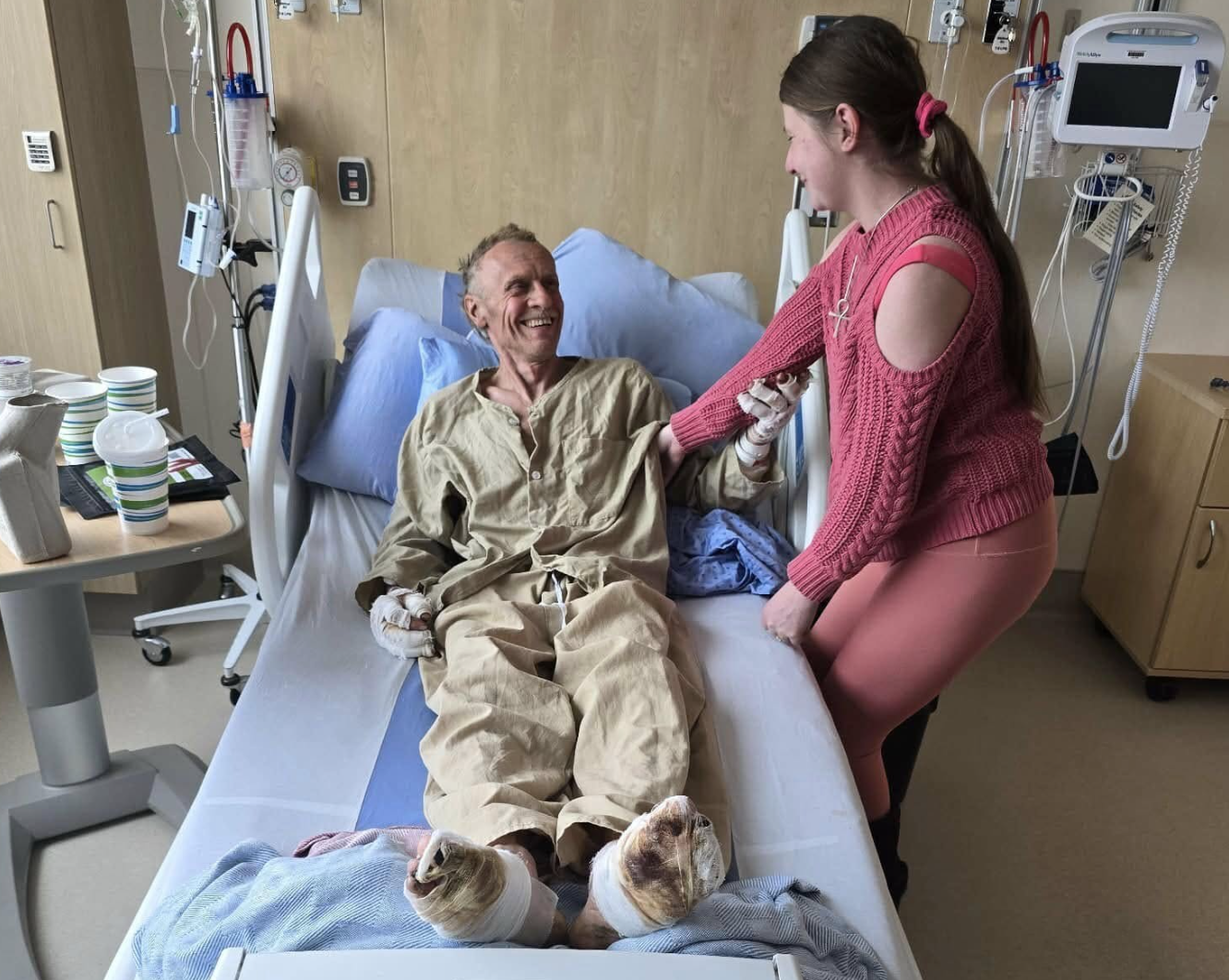 A man smiling in a hospital bed with bandaged feet with a woman standing beside the bed.