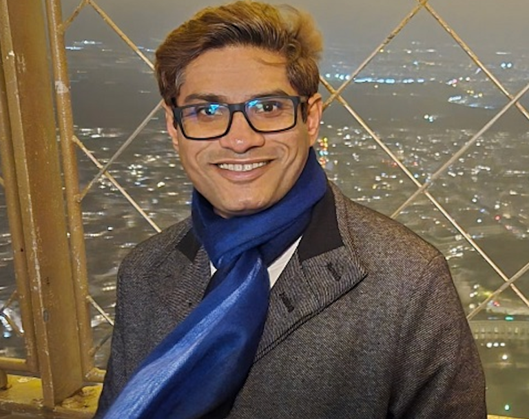 A man in a grey coat, blue scarf and glasses.