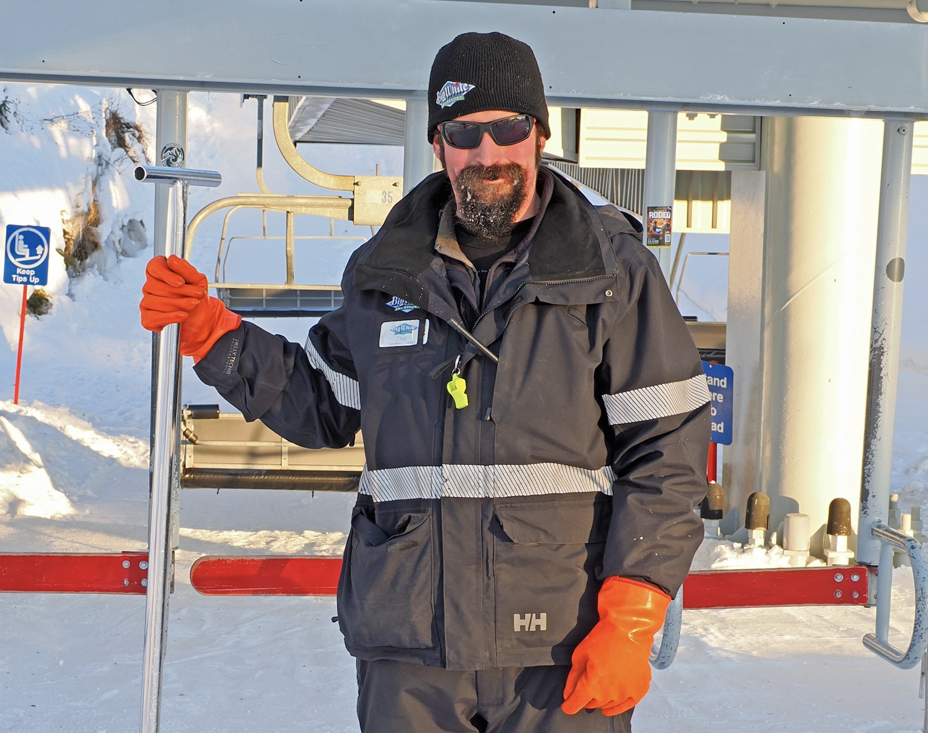 A guy with orange gloves and a rake in front of a ski lift.