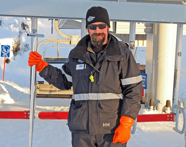 A guy with orange gloves and a rake in front of a ski lift.