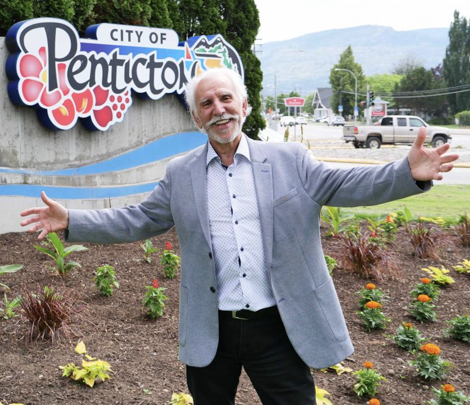 Former Penticton mayor John Vassilaki stands with his arms spread in front of a City of Penticton sign.