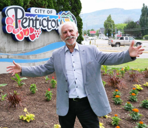 Former Penticton mayor John Vassilaki stands with his arms spread in front of a City of Penticton sign.