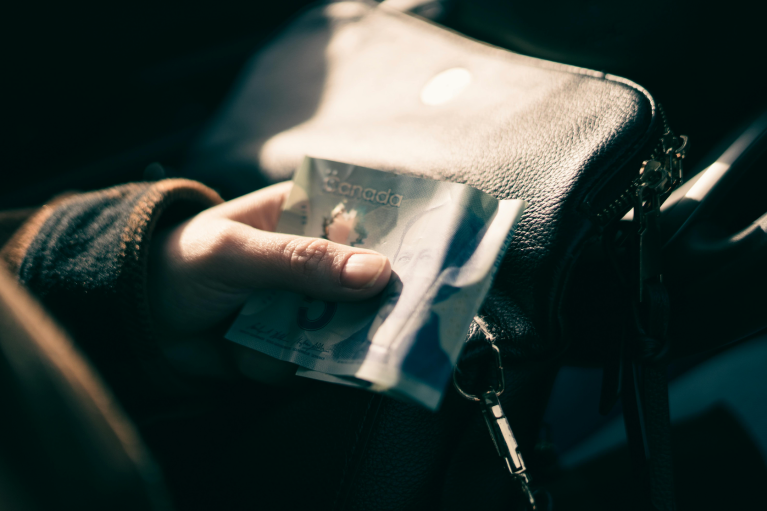 A person holding a change purse, car keys and a five dollar bill against a steering wheel.