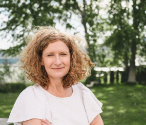 A ginger haired woman wearing a white T-shirt stands in front of trees.