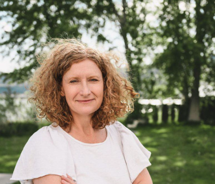 A ginger haired woman wearing a white T-shirt stands in front of trees.