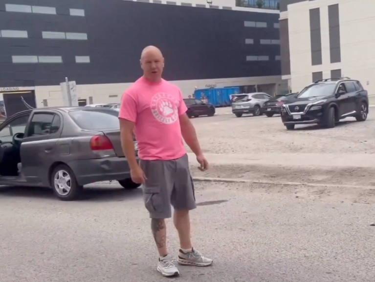 A bald man wearing a pink shirt yells at a person making a video if him.