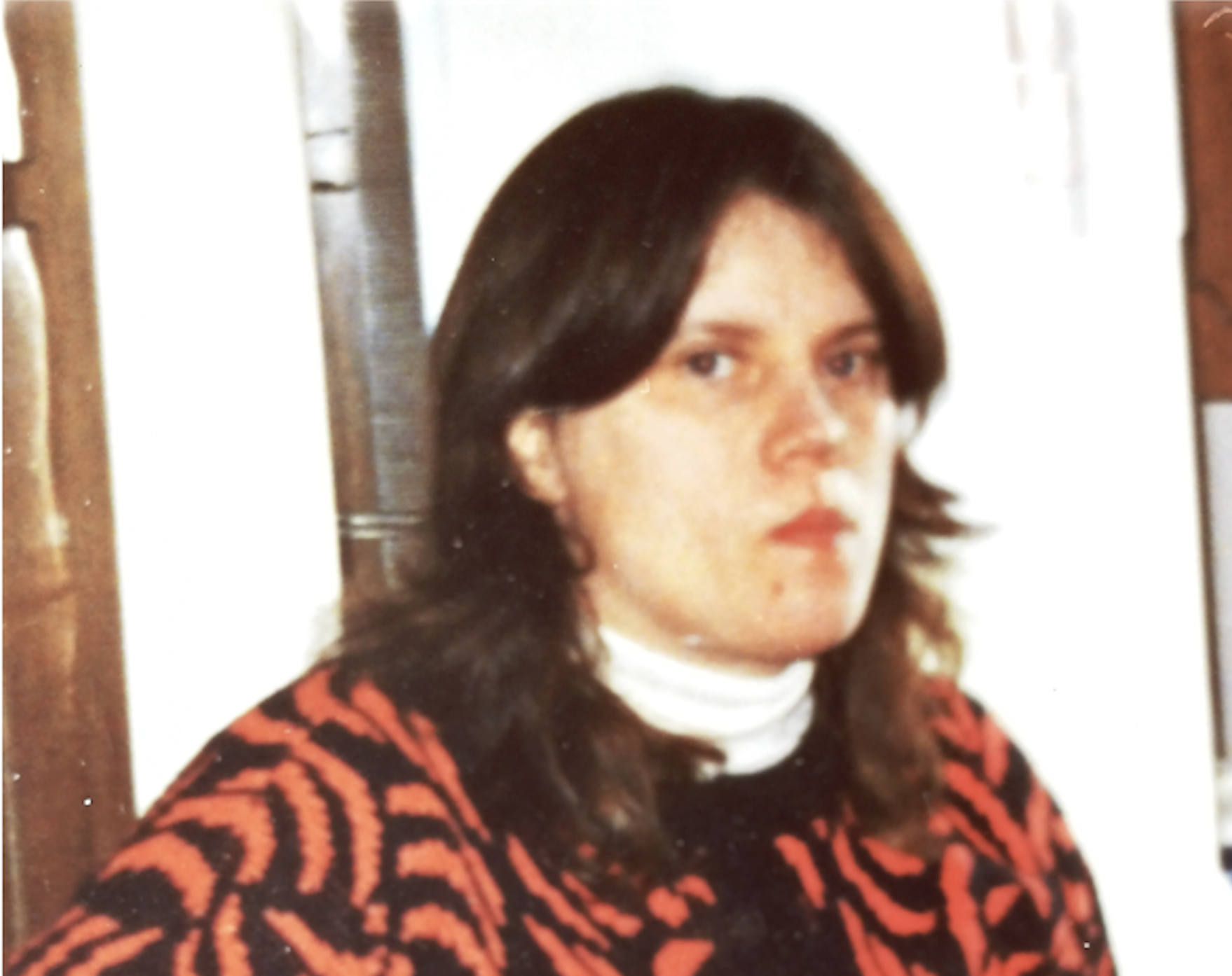 A woman wearing a red and black patterned sweater over a white turtle neck poses for a photo.