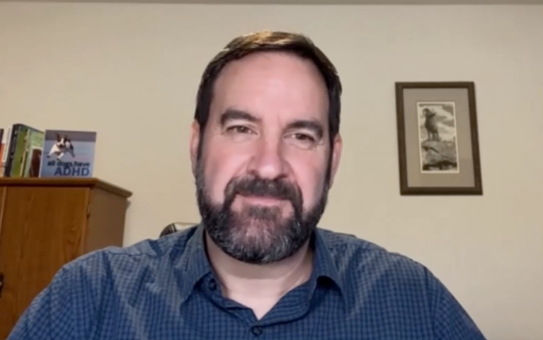 A man with a beard wears a blue shirt in a head and shoulders shot.