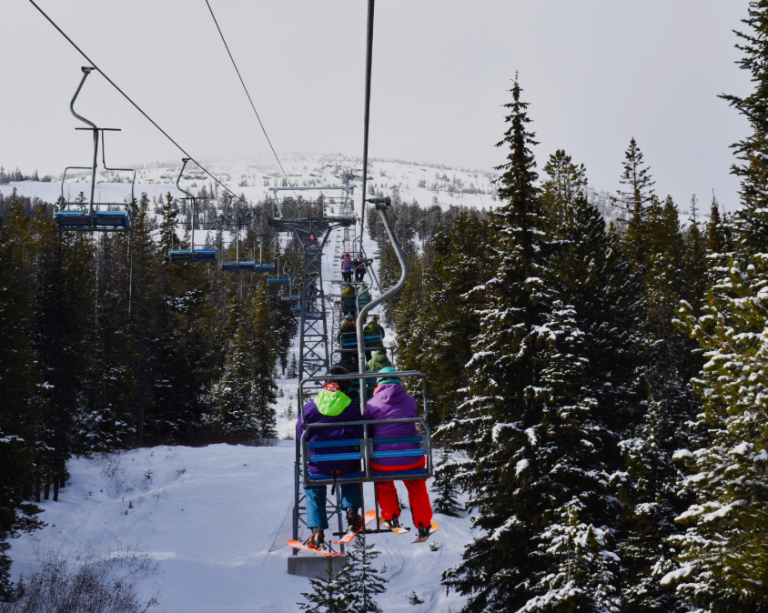 Two people sit on a ski lift, surrounded by tress.